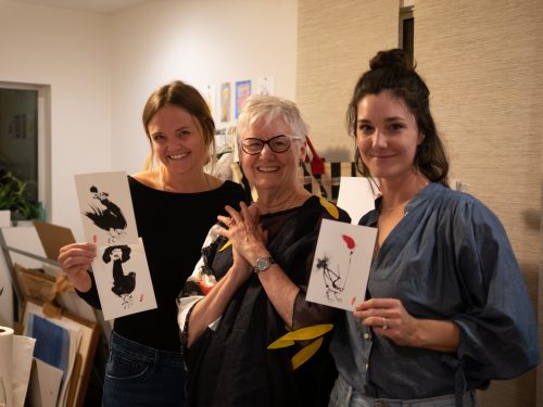 Three women holding up paintings of zen chickens.